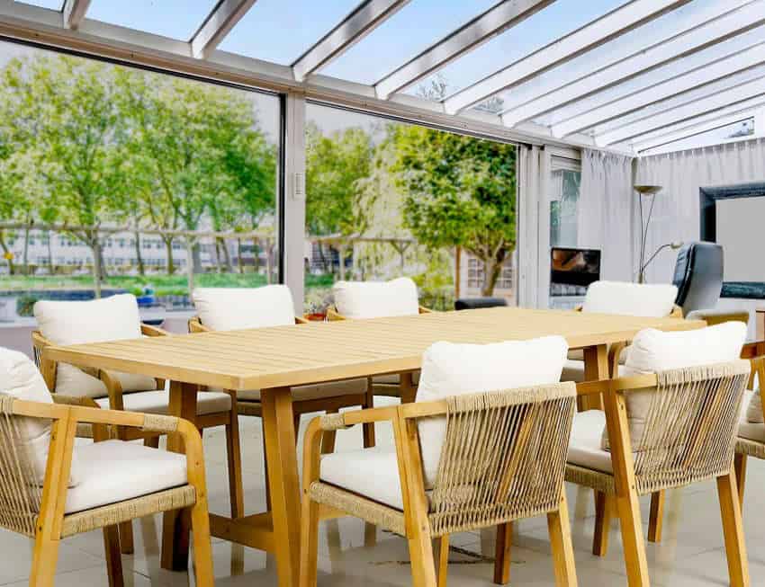 A bright, modern sunroom with a large wooden dining table and eight chairs featuring rope detailing and white cushions, set under a glass ceiling. Lush green trees are visible through the expansive windows.