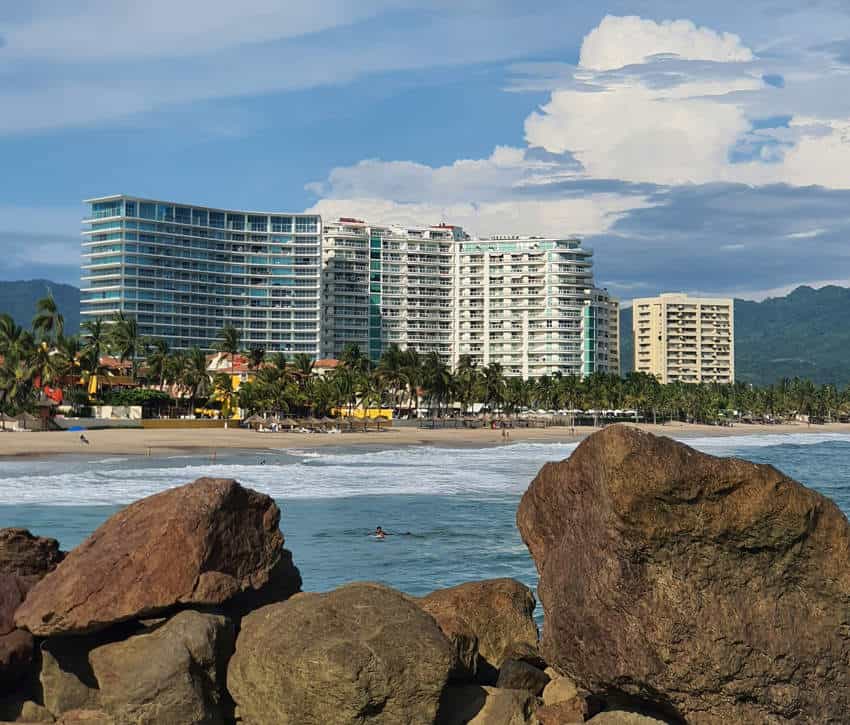 A view of a beach with large, brown rocks in the foreground, and ocean waves gently rolling onto the shore. In the background, modern, multi-story resort buildings line the sandy beach, backed by palm trees and lush green mountains under a partly cloudy sky.