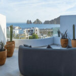 A modern rooftop patio features a curved grey sofa, potted cacti, and a panoramic view of the ocean, a city, and distant rock formations under a clear sky.