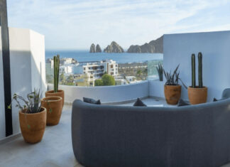 Want to buy property in Mexico? Make sure you invest close enough to a major airport A modern rooftop patio features a curved grey sofa, potted cacti, and a panoramic view of the ocean, a city, and distant rock formations under a clear sky.