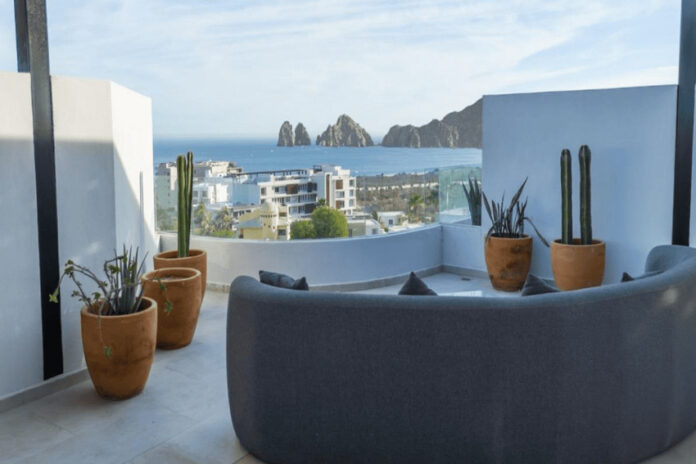 A modern rooftop patio features a curved grey sofa, potted cacti, and a panoramic view of the ocean, a city, and distant rock formations under a clear sky.
