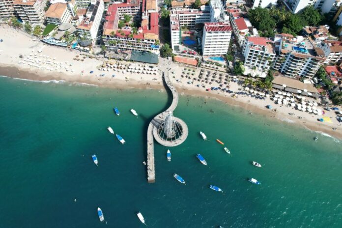 Aerial view of a winding pier with a circular end extending into clear blue-green ocean, with a sandy beach and a city skyline visible beyond.
