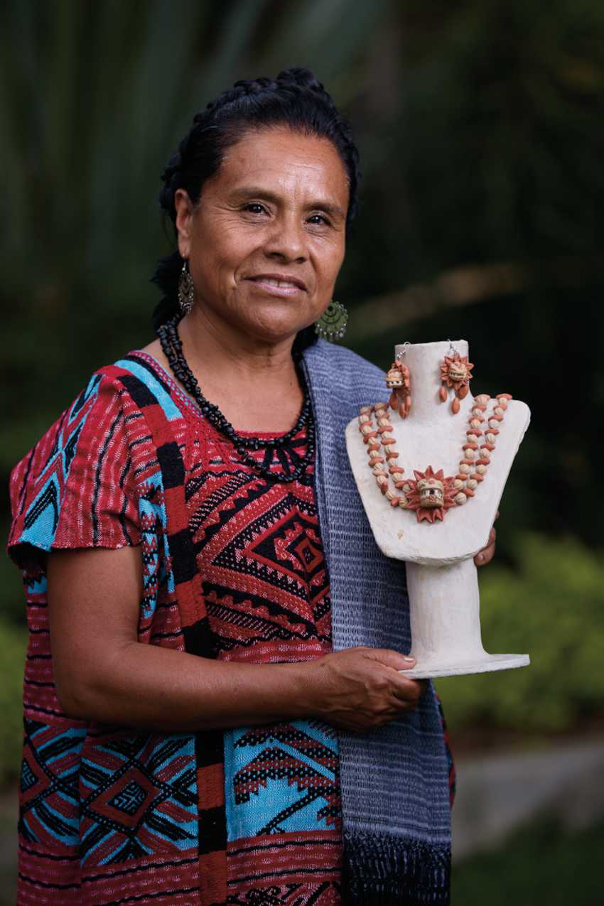 A Mexican woman with dark skin and braided hair, wearing a traditional red and blue Indigenous embroidered dress and a dark beaded necklace, stands smiling while holding a white jewelry display bust featuring a light-colored traditional handmade necklace and matching earrings adorned with small carved faces. A blue shawl is draped over her left shoulder, and a leafy green background is blurred behind her.