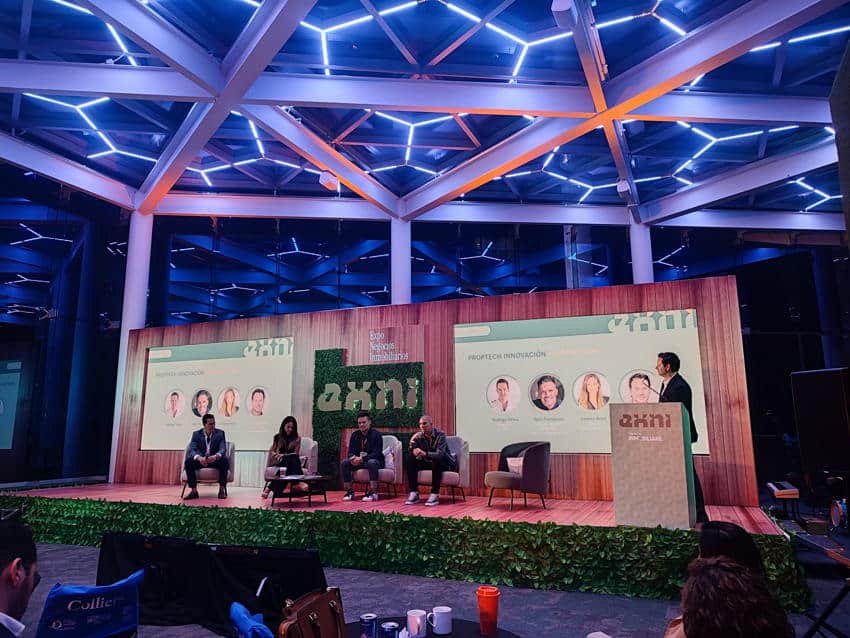 A wide, low-angle shot captures a modern conference stage with a panel discussion in progress, illuminated by a geometric, glowing blue and white ceiling structure. Four panelists are seated in armchairs on a raised wooden platform, facing a large screen displaying "PROPTECH INNOVACIÓN" and images of four individuals. To the right, a presenter in a dark suit stands at a podium with the "EXNI" logo. The stage backdrop features a wooden texture with white horizontal lines and additional "EXNI" branding. The foreground shows the backs of audience members.