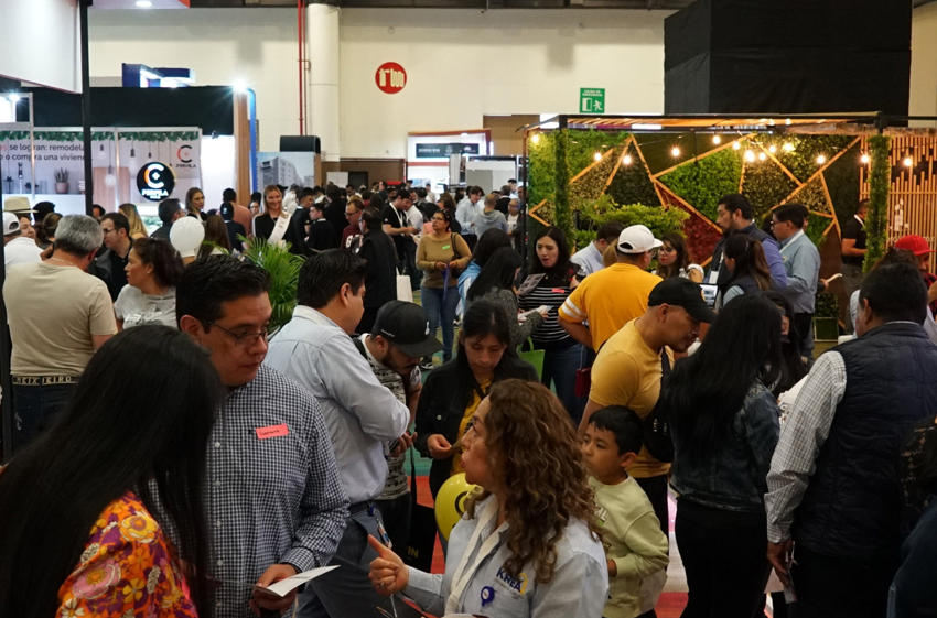 Bustling indoor exhibition hall filled with a diverse crowd of people of all ages. Various booths and displays are visible, including one with a green wall and string lights, and another with hanging light fixtures. Many attendees are standing, mingling, and looking at the exhibits.