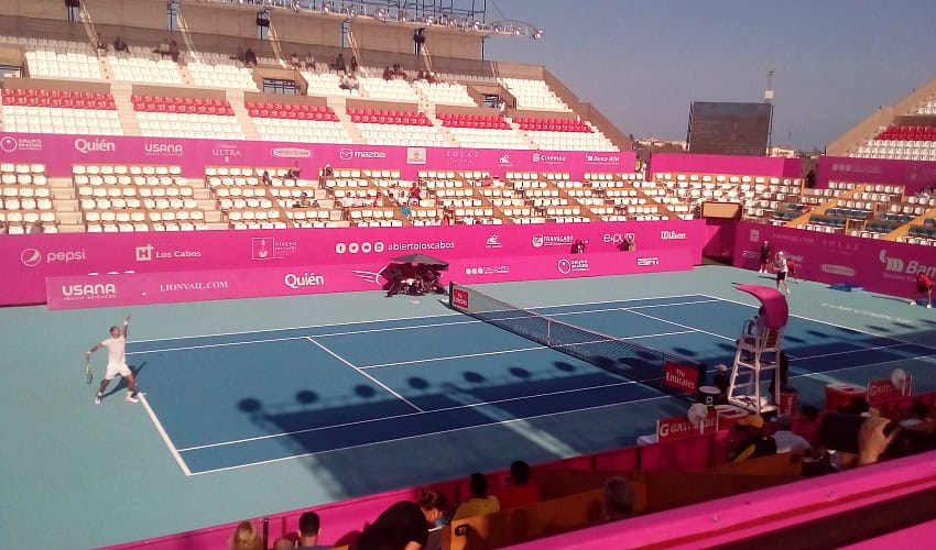 A wide shot of a blue outdoor tennis court during a match, with a player in white serving on the left. The stadium seating, predominantly white with some pink accents, is visible behind the court, largely empty but with a few spectators. Advertising banners in pink line the court, and a chair umpire sits at the net. The sky is bright and clear.