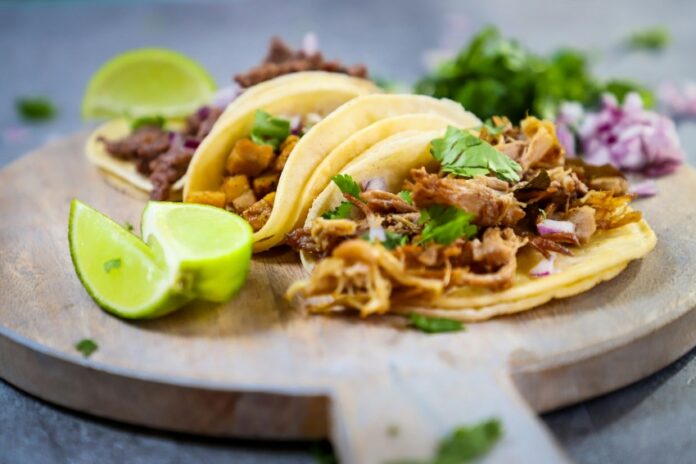 Mexican style tacos filled with meat, red onions and cilantro with some lime wedges on the side, all on a round, wooden charcuterie board.