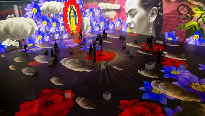An immersive art exhibit with images projected onto the walls and floor. The projections include large blue-and-red flowers, a prominent image of Frida Kahlo's face, a depiction of the Virgin of Guadalupe, and floating white clouds. Several people are scattered throughout the exhibit, observing the art.