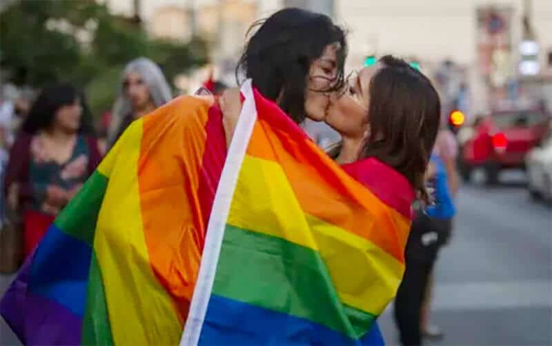 Two women kissing in the street wrapped in a multicolored rainbow gay pride flag.