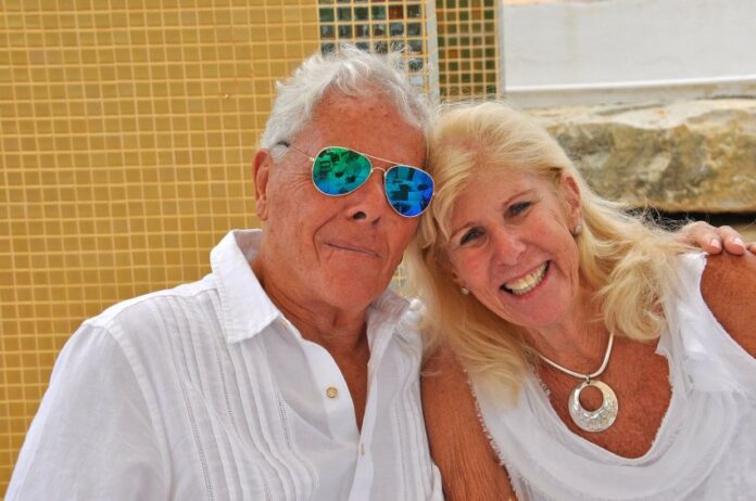 An elderly man and woman holding each other and posing for the camera outside against a Mexican tiled wall.