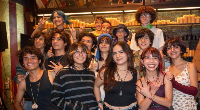 A diverse group of smiling Mexican teenagers, with various hairstyles and clothing styles, pose for a group photo in a dimly lit restaurant or bar with rows of candles in the background.