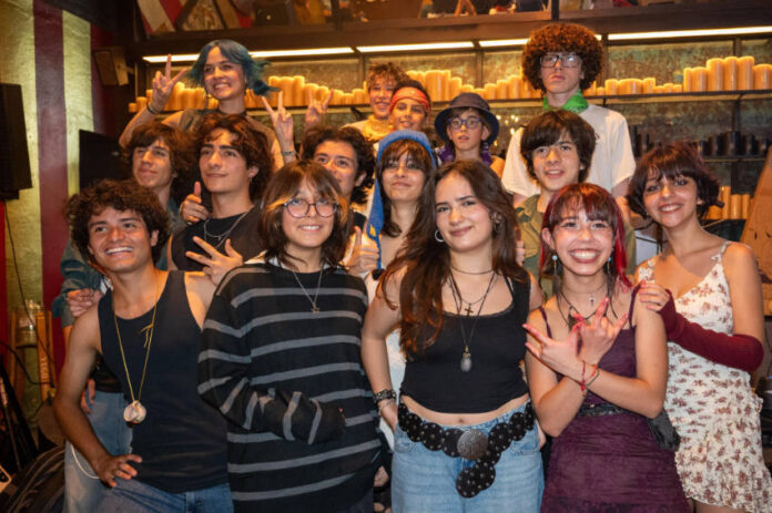 A diverse group of smiling Mexican teenagers, with various hairstyles and clothing styles, pose for a group photo in a dimly lit restaurant or bar with rows of candles in the background.