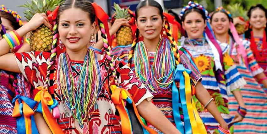 A group of smiling young Mexican women, adorned in vibrant traditional attire of indigenous Oaxaca, participate in a cultural celebration. In the foreground, two women with intricately braided hair decorated with colorful ribbons wear richly embroidered blouses and necklaces made of countless strands of multicolored beads. Some of the women hold a pineapple on their right shoulder. Other women in similar festive clothing are blurred in the background, suggesting a parade or dance.