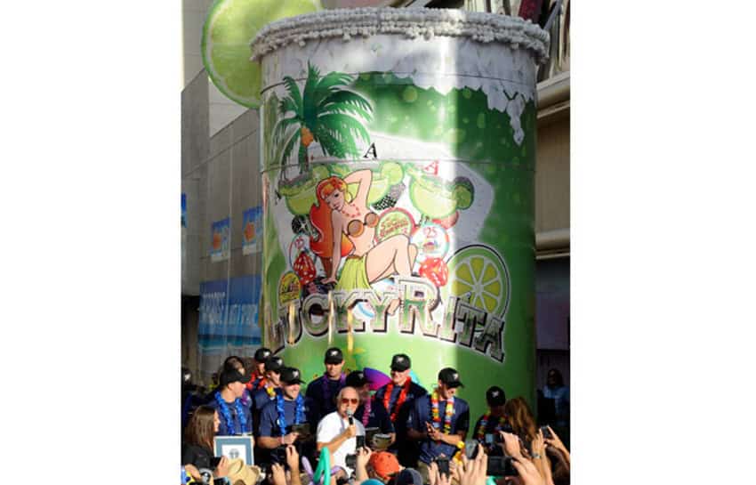 A giant, oversized 'Lucky Rita' margarita glass statue, featuring artwork of a woman in a grass skirt and a bikini top, surrounded by palm trees and limes and margaritas in cocktail glasses. People are standing in front of the statue, one in front center holding a microphone. They are being celebrated by a crowd of people, of which we can only see their upraised hands.