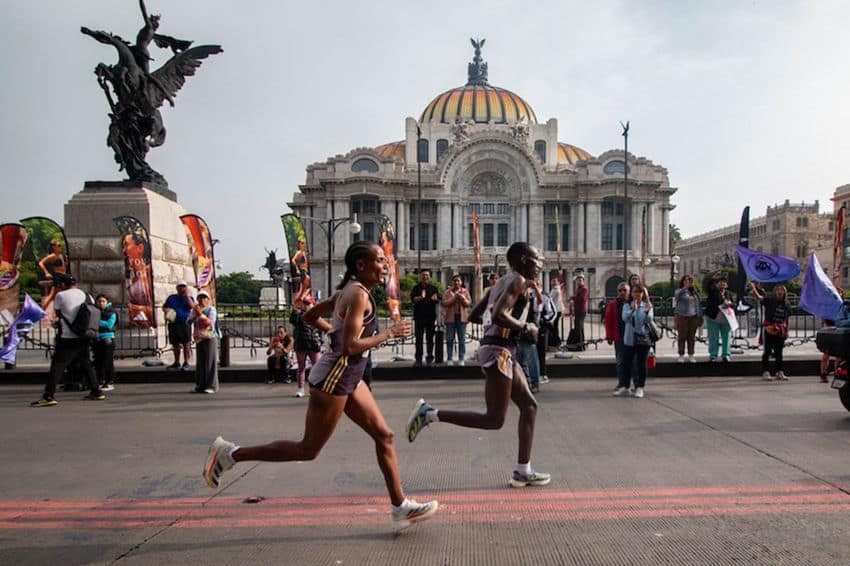 Two runners are captured mid-stride as they race past the iconic Palacio de Bellas Artes in Mexico City. The runners, wearing athletic gear, are crossing what appears to be a designated race route marked on the pavement. Behind them stands the Palacio with its distinctive Art Nouveau and Art Deco architecture, featuring a prominent golden dome and ornate facade. To the left of the frame is a bronze monument with winged figures. The scene is bustling with spectators, race officials, and colorful banners