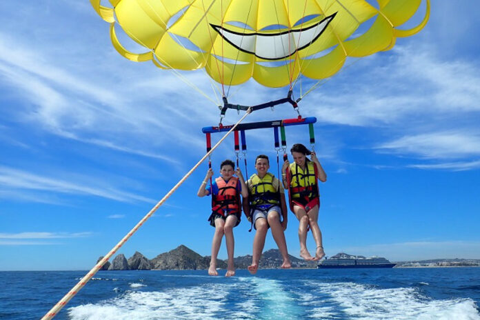 Three people strapped into a yellow parasail connected to a boat off camera. They are in the air slightly above the ocean.