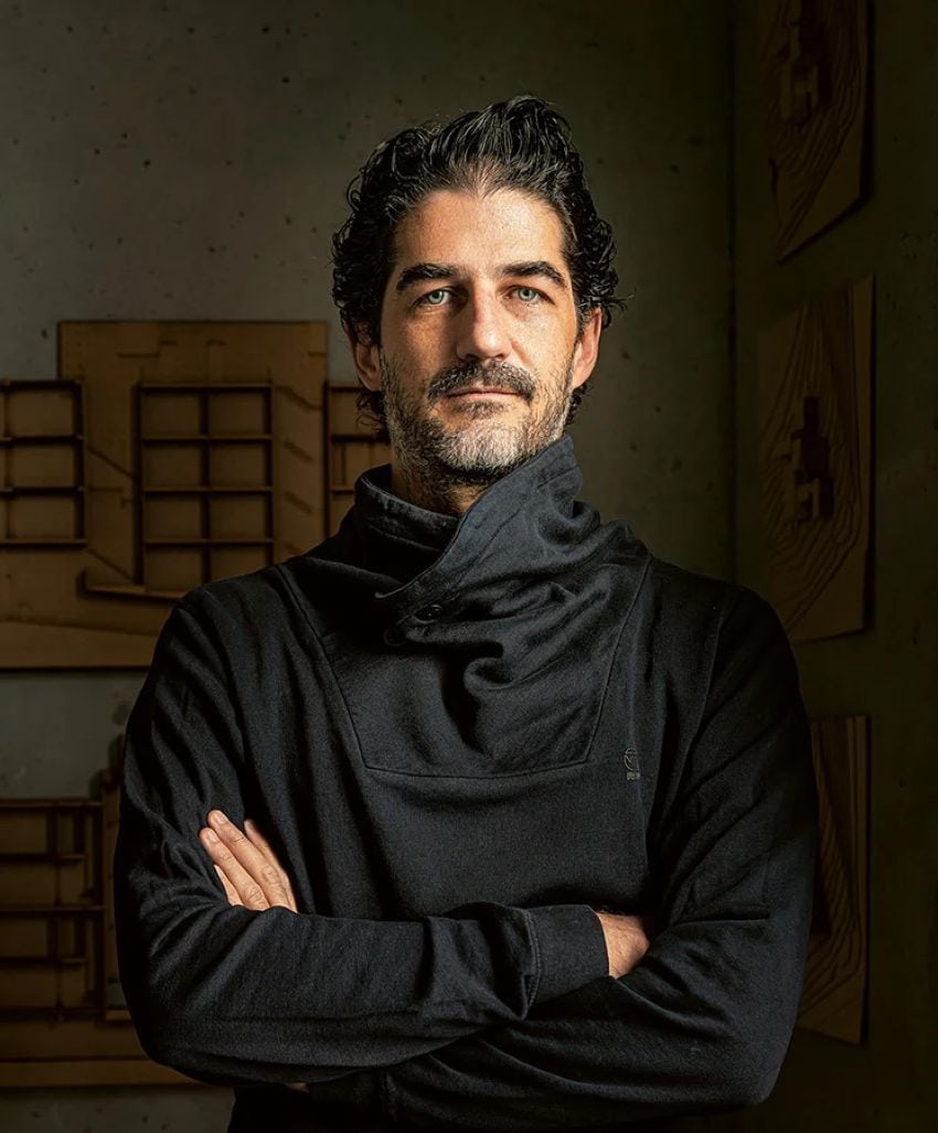 Mexican architect Hector Barroso posing for a publicity photo in a shadowy room. He is staring into the camera with his arms crossed, dressed all in black.