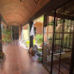 A colonial-style covered corridor with terracotta tile floors, arched brick ceilings, and large windows overlooking a courtyard garden in San Miguel de Allende.