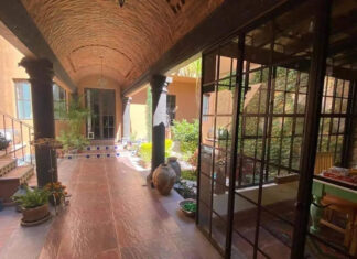 A colonial-style covered corridor with terracotta tile floors, arched brick ceilings, and large windows overlooking a courtyard garden in San Miguel de Allende.