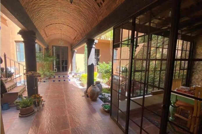 A colonial-style covered corridor with terracotta tile floors, arched brick ceilings, and large windows overlooking a courtyard garden in San Miguel de Allende.