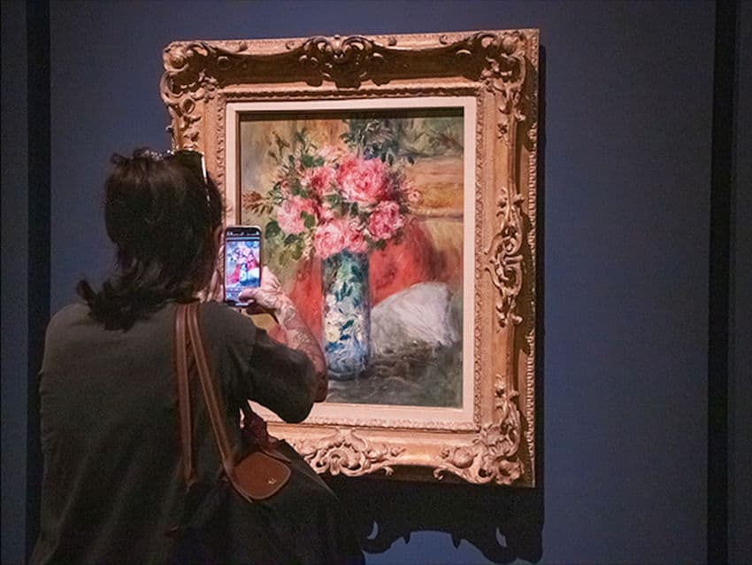 A person with their back to the camera takes a photo of a painting of pink flowers in a vase at an art exhibition. The painting is in an ornate gold frame and depicts a bouquet of pink roses and other flowers in a decorative blue and white vase, set against a warm, reddish background.