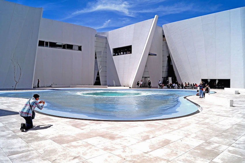 A man kneels to take a photo in a massive courtyard outside a concrete building designed in triangular polygons. In the courtyard's middle is a shallow pond structure made to look like a whirlpool.