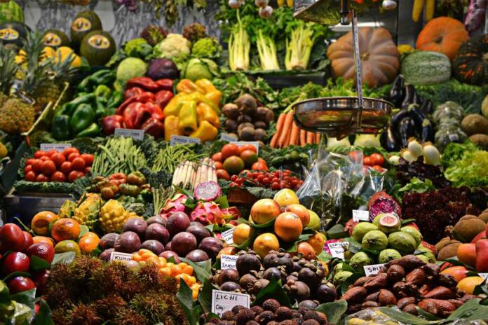 An open air market in a Spanish-speaking country where a wide variety of fruits and vegetables are on display with their prices. In the foreground, an old-fashioned hanging scale for food items can be seen.