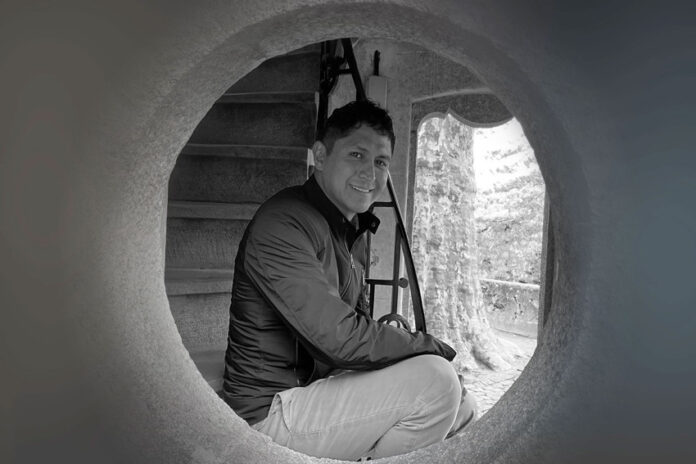 Black and white photo of artist Jaime Colín, a man with short dark hair, smiling and looking directly at the camera. He is crouched inside a large circular opening, with a staircase visible behind him and natural light illuminating a tree-like structure outside the opening.