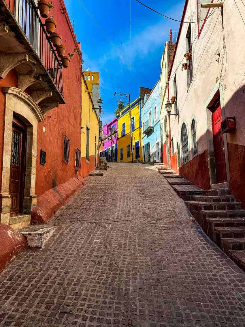 A cobblestoned street going up hill alongside brightly colored antique houses made of stone with old fashioned wooden doors, and decorative balconies
