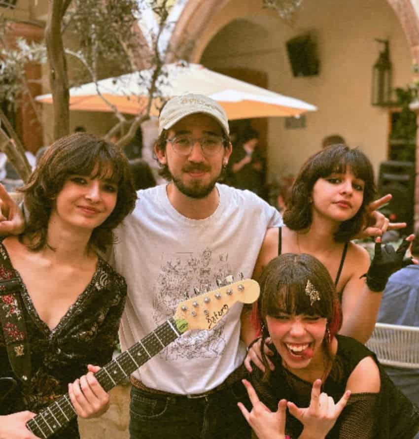 A young man and three young teen girls, who appear to be a band, pose for a photo in an outdoor courtyard. The man, wearing a cap and glasses, has his arms around two of the girls. One holds an electric guitar, while another in the foreground makes a rock-and-roll hand gesture and laughs.