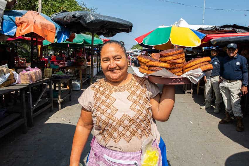 A smiling Oaxacan woman from Juchitan in a patterned blouse and apron holds a tray piled high with golden-brown fritters in an outdoor market with colorful umbrellas in he background.