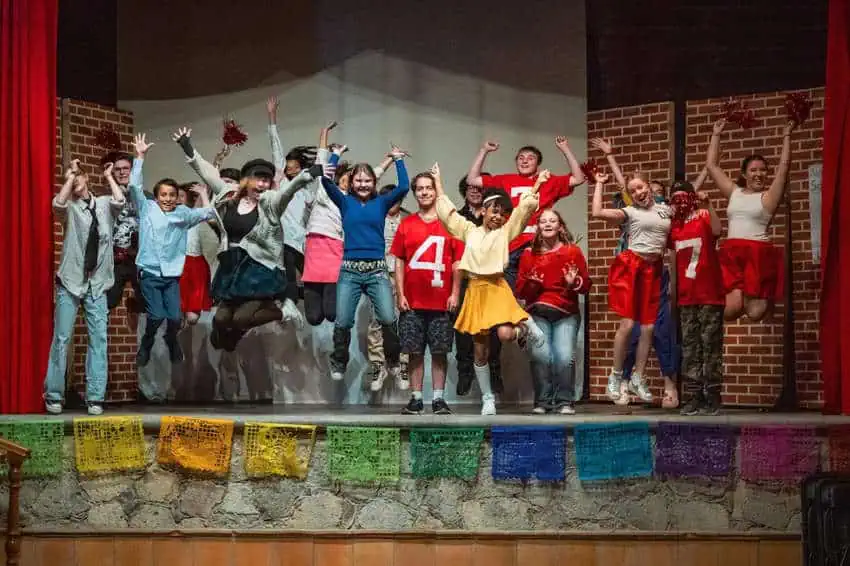 A diverse group of teenagers and a few adults leap joyfully on a stage. Some wave pom-poms, and several wear red and white casual outfits, ranging from jeans to shorts. Colorful papel picado hangs along the front of the stage.