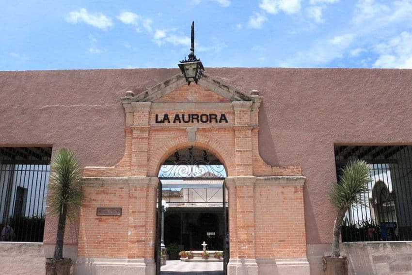 A large brick factory building entrance with the name "La Aurora" in black iron letters up top. There a large windows on either side with wrought-iron bars instead of glass.