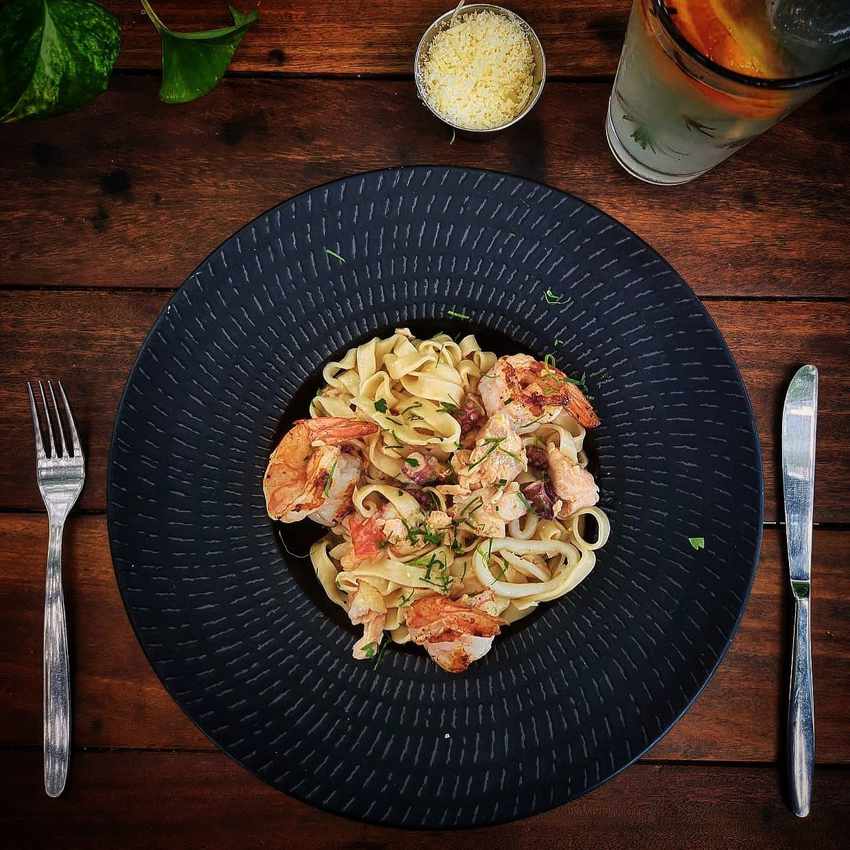 A plate of delicious seafood fettuccine pasta with large shrimp, served on a black textured plate on a wooden table, alongside a glass of fruit-infused water and a small bowl of grated cheese, in a Bacalar, Mexico restaurant.