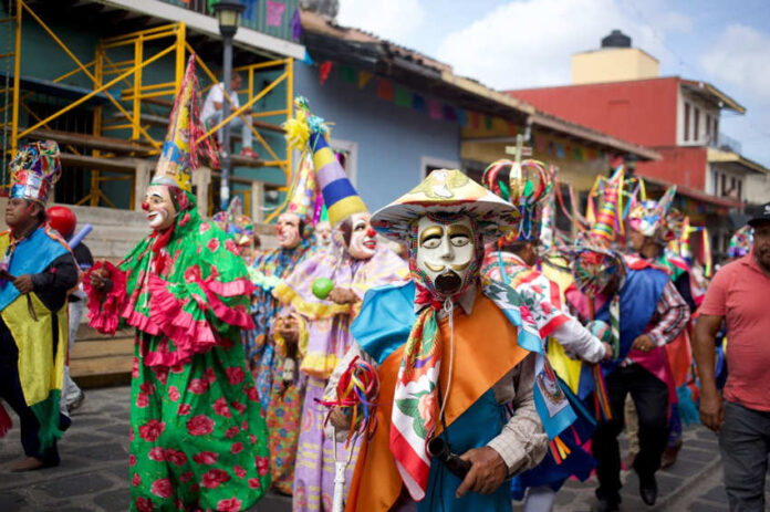 Costumed dancers in vibrant masks and traditional clothing parade through the streets during a Mexican festival in Xico, Veracruz, with colorful banners and colonial buildings in the background.