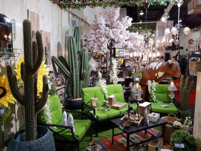 A vibrant Mexican-themed home decor display featuring large artificial cacti, bright green patio furniture, sunflowers, a cow statue, and various rustic lanterns and lighting fixtures, suggesting an eclectic outdoor living space.