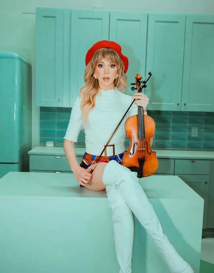 American violinist Lindsay Stirling poses with her violin in a retro-styled kitchen setting, wearing a red beret and white outfit