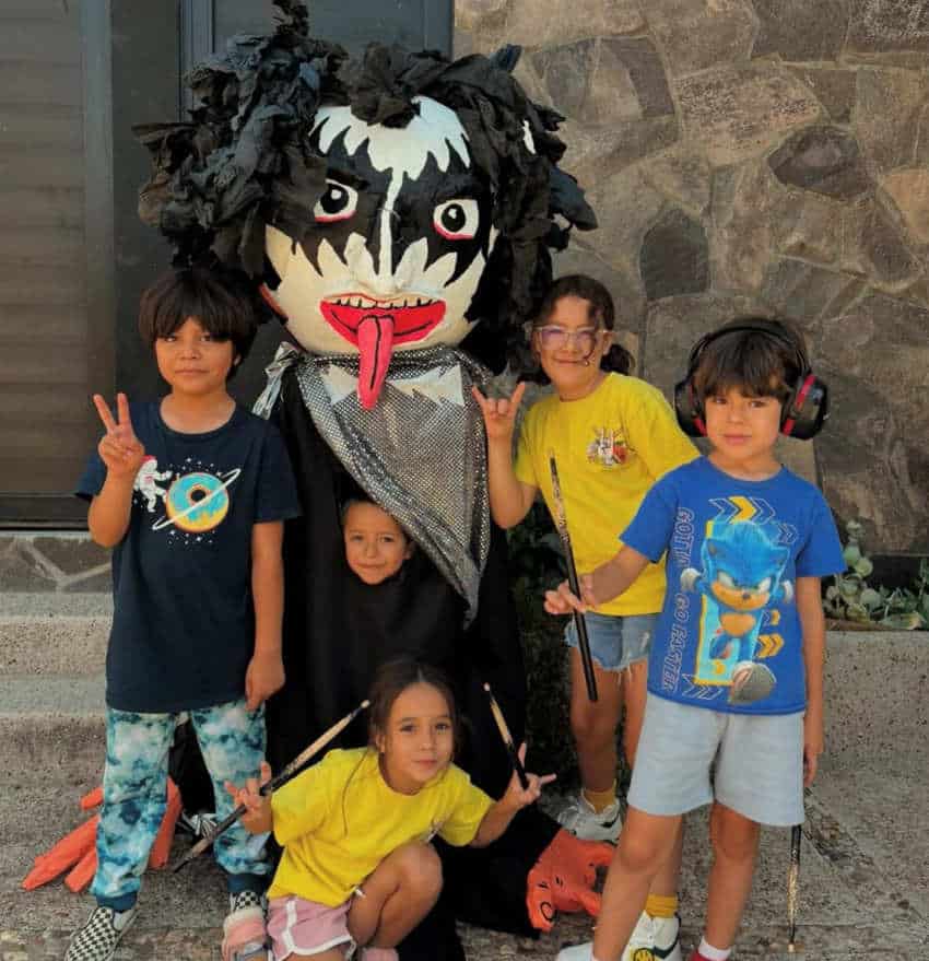 Five young children pose outdoors in front of a stone wall with a large Mexican mojiganga puppet made to look like a member of the rock band KISS in black-and-white face makeup and a long tongue. The children are smiling around it, some holding drumsticks and others making peace or "rock-on" hand signs.