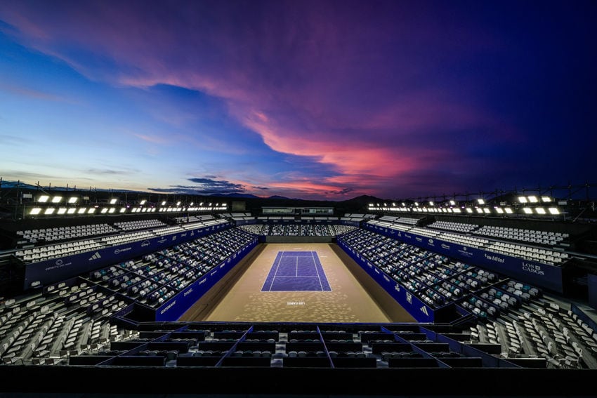 An expansive view of an outdoor tennis stadium at dusk. The hard court is illuminated by bright overhead lights, appearing beige with a dark blue playing area. The stands, filled with rows of empty seats, rise up on either side. A vibrant sunset with hues of pink, purple, and blue fills the sky above distant hills.