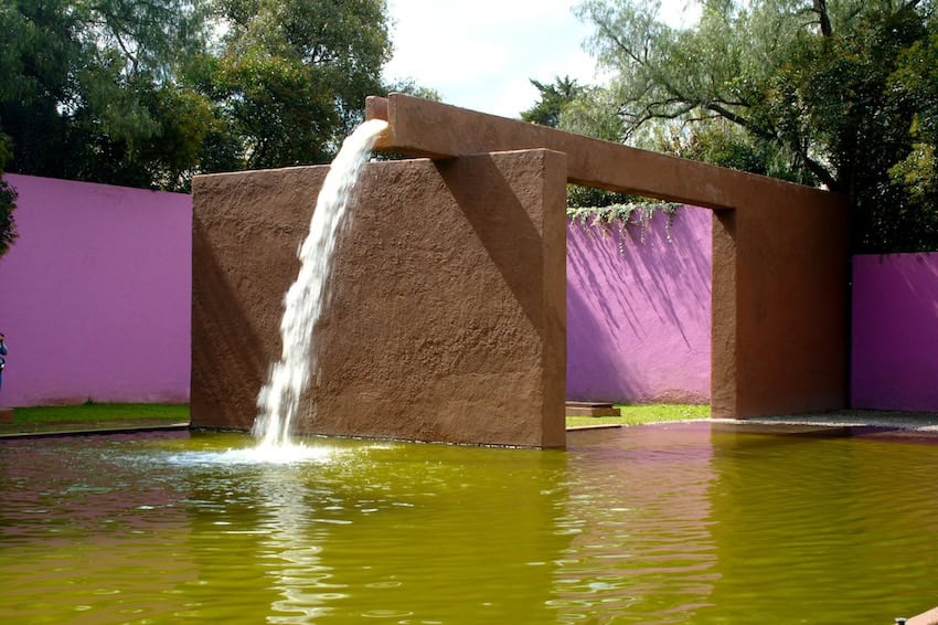 A courtyard in pink and brown painted concrete features two walls that support a an aqueduct allowing water to fall into the courtyard's pool.
