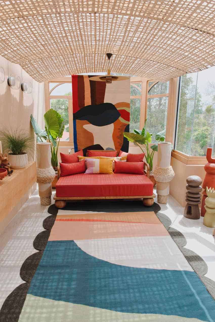 A vibrantly decorated living space in Mexico featuring a red daybed with colorful pillows, a large abstract tapestry on the back wall, and a long, multi-patterned rug on the floor. The room has a woven ceiling, large windows, and is adorned with various decorative vases and plants.
