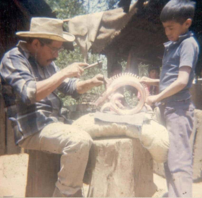 Jesus Perez Ornelas sitting on a tree stump in his workshop in Santa Clara del Cobre, Michoacan, working with a small hammer on pounding the copper of an artisan piece. A 10-year-old Mexican boy holds the piece in place for Perez.