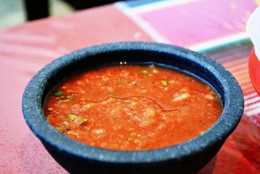 A volcanic-stone bowl filled with thick, red, Mexican-style salsa on a table. The background is in bokeh.