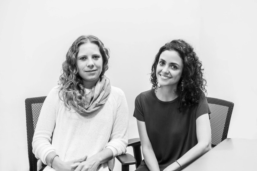 Mariana Ordóñez and Jesica Amescua sitting in executive desk chairs side by side posing for the camera in a completely white room with no features in the background