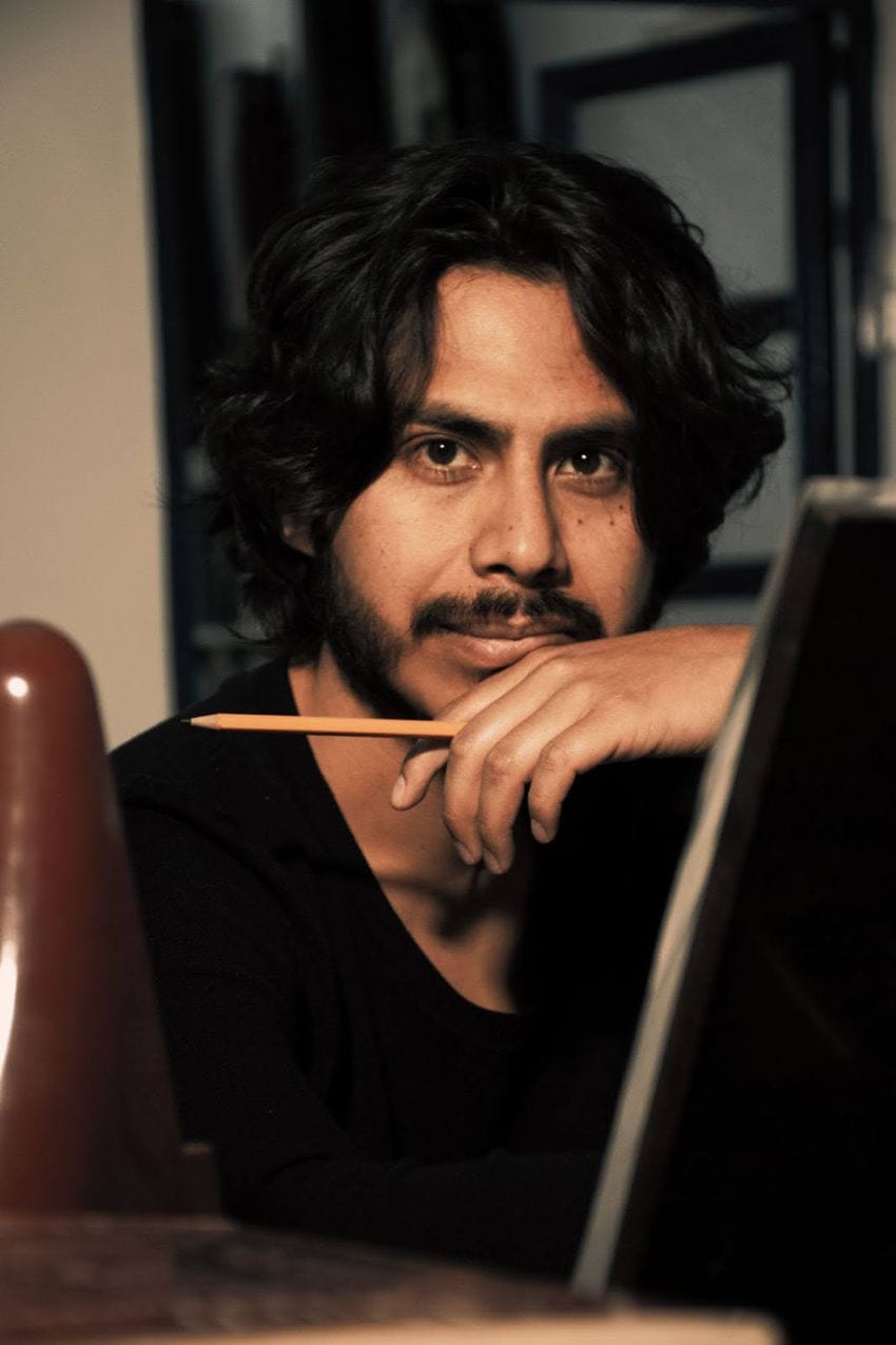 A man with dark wavy hair and a goatee looks thoughtfully at the camera. He is resting his chin on his hand and holding a yellow pencil, seated behind an easel in a warmly lit, artistic setting.