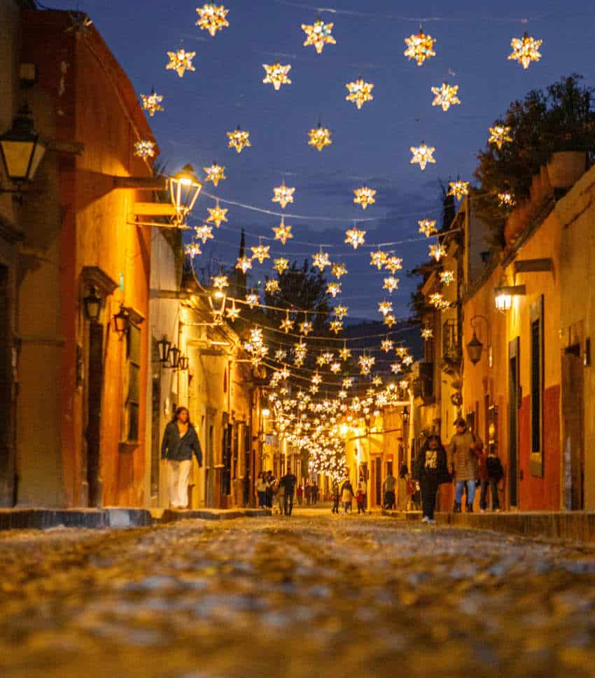 A vibrant, low-angle shot of a narrow cobblestone street in a Mexican town at dusk, illuminated by numerous golden star-shaped lights strung overhead between colorful buildings, with people strolling along the street.
