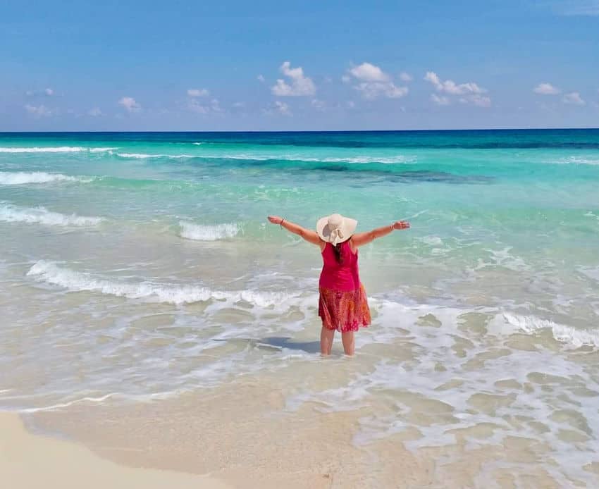 A woman in a sun hat and red dress stands in the clear, turquoise water of a Mexican beach, arms outstretched looking out at the ocean's vanishing skyline