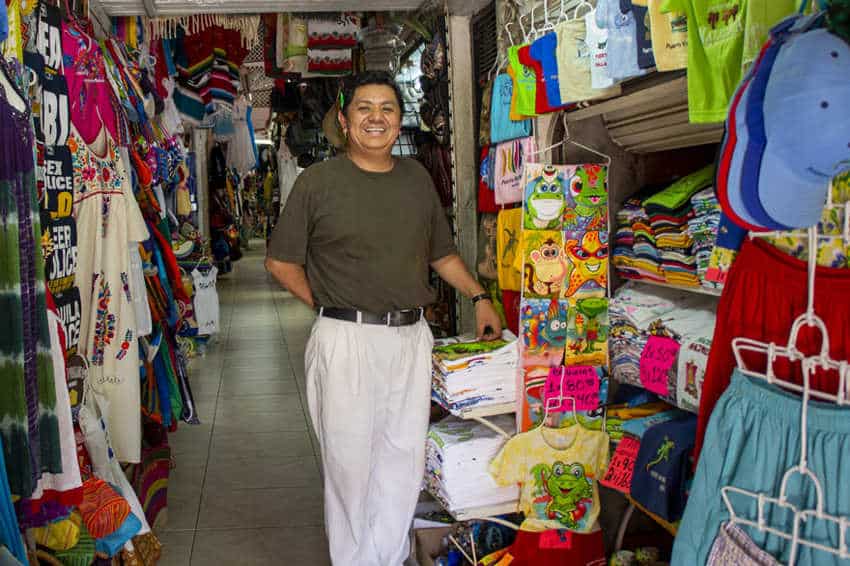 A smiling male vendor stands in his narrow market stall filled with colorful clothing and souvenirs in Puerto Vallarta, Mexico