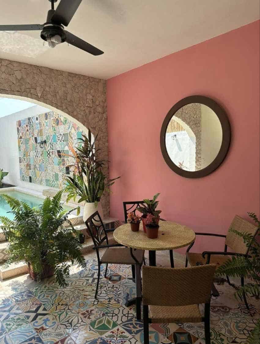 An inviting patio features colorful mosaic tiles on the floor, a pink wall with a round mirror, a small table and chairs, and an arched doorway leading to a pool area with a mosaic-tiled wall.