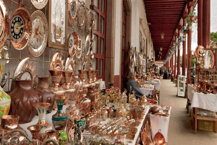 An artisan fair in a Mexican courtyard in a small town where hundreds of coppersmithed decorative objects are laid out on tables.